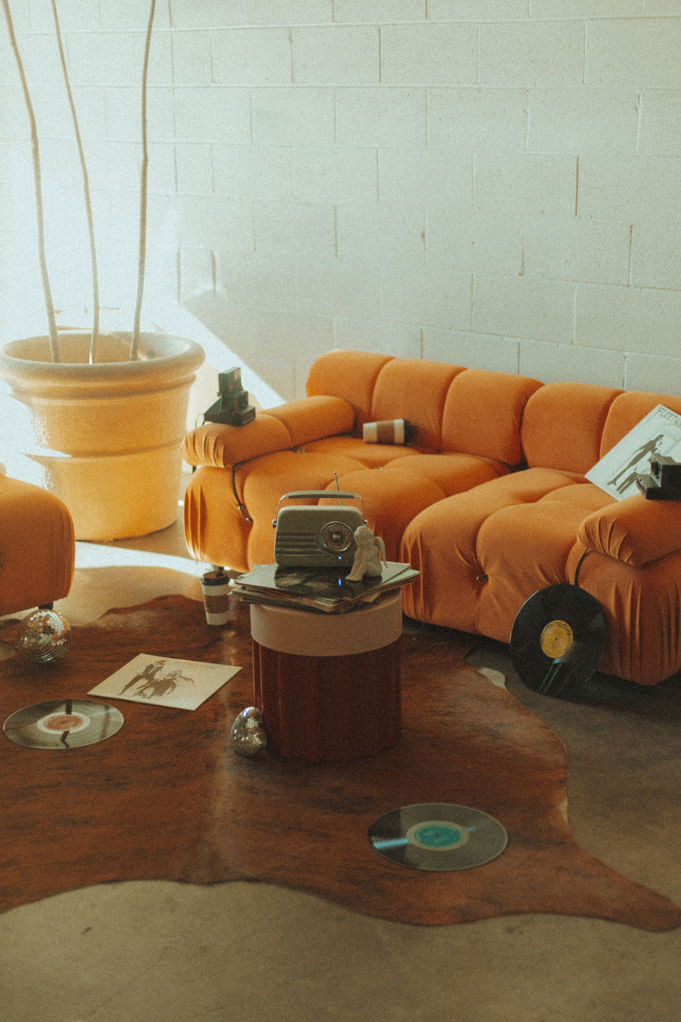 Cozy room with an orange sofa and vintage decor. A retro projector sits on a table surrounded by records on a wooden floor, under soft, warm light.