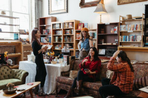 A group of four women enjoy a casual gathering in a cozy bookstore setting. Three are seated on a floral couch, laughing, while one stands nearby, engaging them in conversation. Shelves filled with books line the walls, and a table with snacks and drinks adds to the inviting, joyful atmosphere.