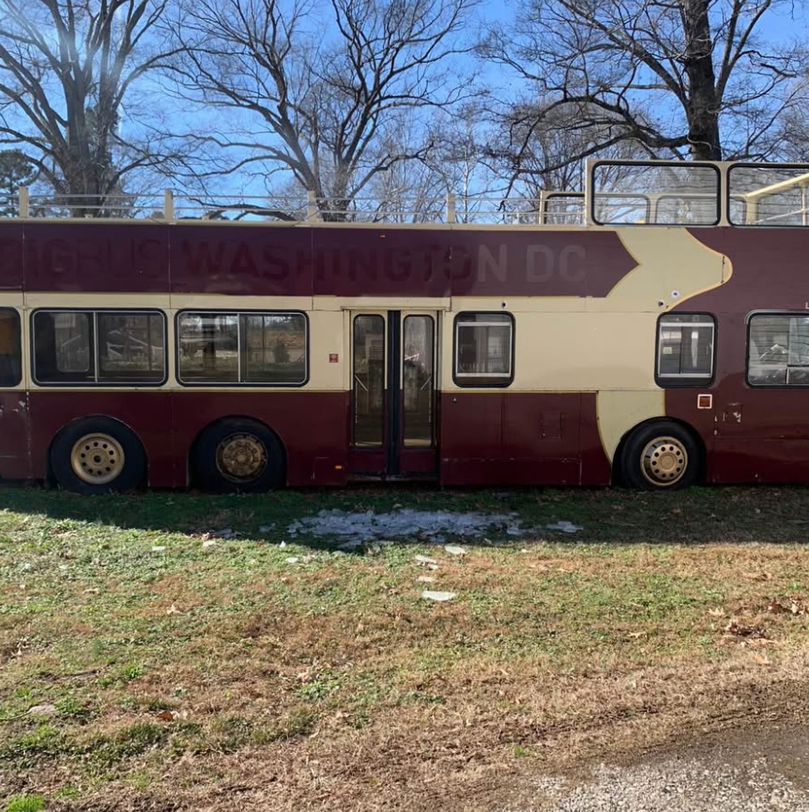 A stationary maroon and cream double-decker bus on grass under leafless trees and a clear sky. The scene suggests abandonment and stillness.
